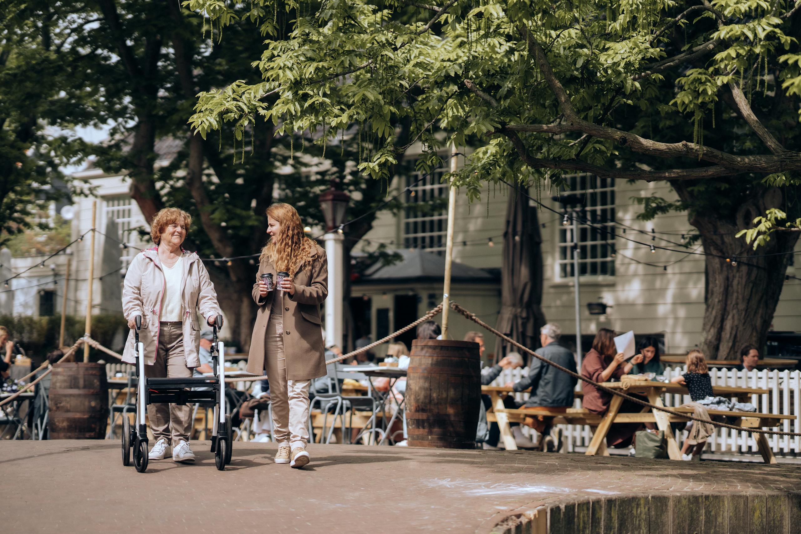 Elderly woman with walker and young companion strolling in a lively Amsterdam neighborhood.